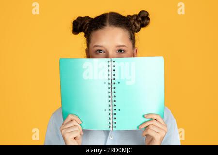 Lächelndes europäisches, kluges Mädchen bedeckt Gesicht mit Buch, bereite dich auf den Test vor Stockfoto