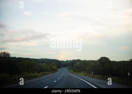 Eine Landschaft mit einem Anwesen am See, umgeben von Wäldern, die die Straße hinunter fahren, mit einem Regenbogen am Ende und Regentropfen auf der Fensterbank. Stockfoto