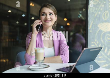 Eine lächelnde Geschäftsfrau, die mit Kunden oder Geschäftspartnern telefoniert, während sie in einem Café mit Kaffee und Paptop sitzt Stockfoto