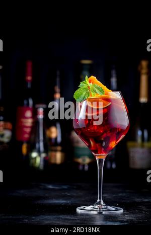 Rotes Sangria-Sommergetränk mit spanischem Rotwein, Pfirsich, Pflaume, Orange, Limette und Eis. Schwarzer Bartheke-Hintergrund, Stahlbarren und Flaschen Stockfoto