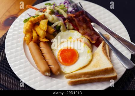 Frühstück mit Würstchen, Eiern, Brot, Speck, pommes frites und frischem Salat in einem weißen Gericht auf einem Holztisch, Bild von Speisen aus nächster Nähe. Stockfoto