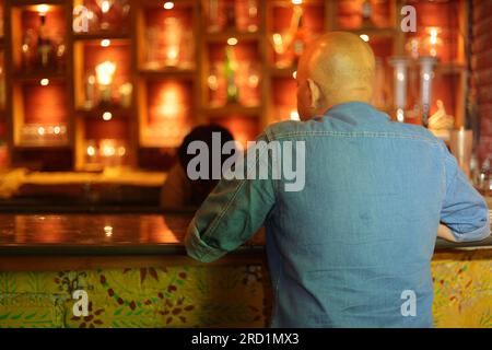 Ein junger und extravaganter, gutaussehender, glatzköpfiger Kerl, der von hinten an der Theke erschossen wurde. Ein attraktiver Mann, der im Nachtklub sitzt. Schnaps bestellen. Stockfoto