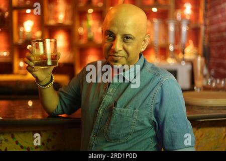 Dramatische Aufnahme eines jungen und attraktiven, glatzköpfigen Kerls, der an der Theke steht. Genießen Sie die Inneneinrichtung des City Clubs im Nachtklub mit einem Glas Whiskey Stockfoto