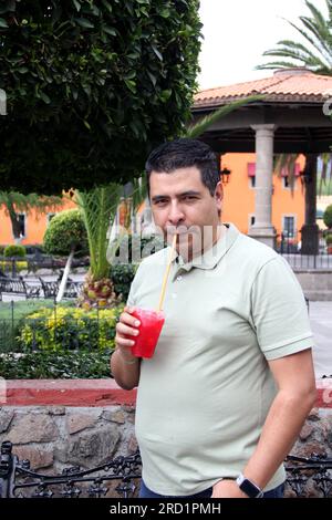 Der dunkelhaarige Erwachsene Latino-Mann macht eine Pause und entspannt sich auf einem Spaziergang im Park, der mit Johannisbeeren aromatisiertes, rasiertes Eis trinkt Stockfoto