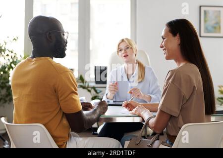 Rückansicht eines multiethnischen jungen Paares, das einen Heiratsantrag stellt, während er mit einem Sozialarbeiter am Tisch sitzt Stockfoto