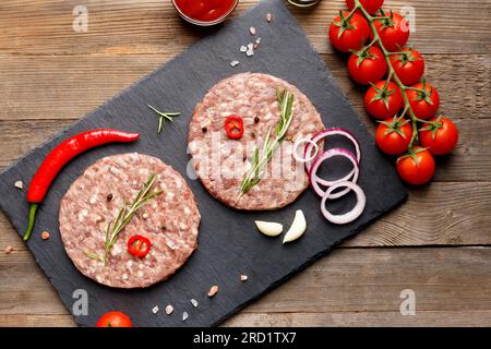 Rohe, frisch gemahlene Rinderburger, Rosmarin, Gemüse und Gewürze auf einer schwarzen Steinplatte auf Holzhintergrund, Draufsicht. Stockfoto