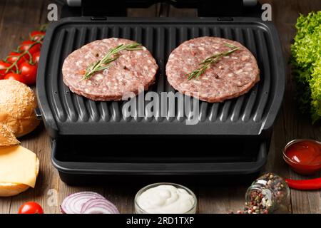 Rinderpasteten für einen Burger werden auf einem eleganten elektrischen Grill auf rustikalem Holzhintergrund gebraten. Hausgemachte Hamburger. Zutaten für Burger. Stockfoto