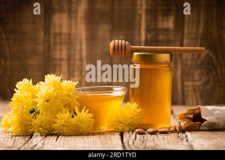 Honig in einem geschlossenen Glasgefäß und einer Schüssel, Stäbchen für Honig und verstreute Mandeln auf einem hölzernen Hintergrund. Zusammensetzung von Honigglas, Pendelarm, Nüssen in einem Beutel Stockfoto