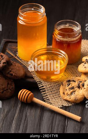 Honig in offenen Gläsern und in einer Schüssel, Dipper und hausgemachte Kekse mit Schokoladenstückchen auf Holzhintergrund. Hausgemachte Kuchen und Honig zum Frühstück. Stockfoto