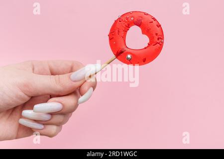 Weibliche Hand mit herzförmigem Lutscher auf pinkfarbenem Hintergrund. Romantische Wohnung zum Valentinstag, Blick von oben Stockfoto