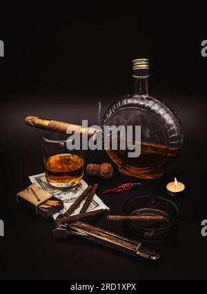 Hochwertige Zigarren mit Cognac, Cutter, 100-Dollar-Papier, Aschenbecher, heller Kerze und rotem heißem Pfeffer auf schwarzem Hintergrund Stockfoto