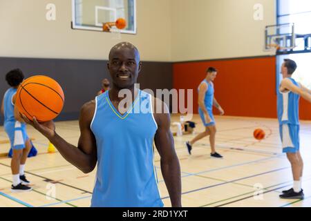 Porträt eines glücklichen afroamerikanischen Basketballspielers mit Basketball über Teamkollegen im Fitnessstudio Stockfoto