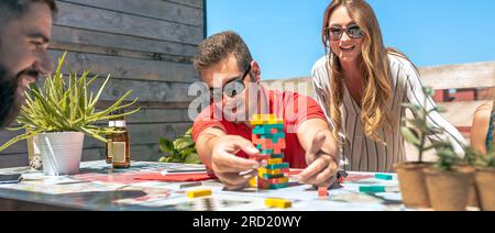 Konzentrierter junger Mann mit Sonnenbrille, der Jenga-Spielchen neben seinen glücklichen Bürokollegen in einer Kaffeepause auf der Terrasse fängt Stockfoto