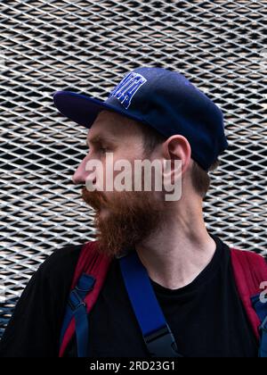 Profilporträt eines bärtigen Millennials-Hipster-Mannes im Snapback auf Metallzäunen-urbanen Hintergrund. Stylischer, selbstbewusster, authentischer Kerl mit rotem Bart und lässiger Freizeitkleidung Stockfoto