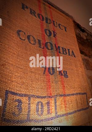 Warm beleuchteter Sackleinen-Kaffeesack mit der Aufschrift „Produkt aus Kolumbien – 70 kg“, der traditionelle Verpackungen zeigt, die im weltweiten Kaffeehandel verwendet werden Stockfoto