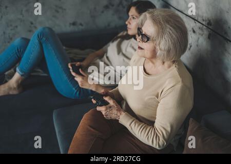 Frau mit ihrer Großmutter, die Videospiele mit dem Controller oder Joystick spielt, während sie auf dem Sofa sitzt. Spiele auf der Konsole. Sie spielen kabellos Stockfoto