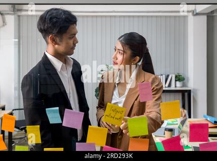 Ein professioneller Mann und eine professionelle Frau stehen zusammen, nehmen an einer Brainstorming-Sitzung Teil und verwenden farbenfrohe Haftnotizen auf einer durchsichtigen Glaswand. Stockfoto