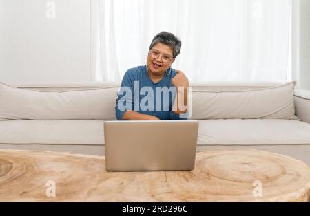 Ältere Frau, die bequem auf dem beigen Sofa saß und an ihrem Laptop arbeitete. Sie ist von einem gemütlichen Wohnzimmer umgeben. Stockfoto