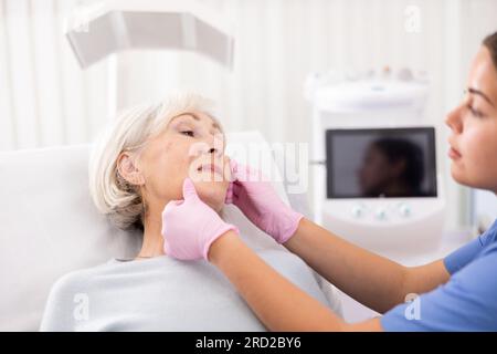 Reife Frau, die während der Beratung mit der Kosmetikerin auf der Couch liegt, ihre Haut vor einem medizinischen Eingriff in der Asthetik-Klinik überprüfen Stockfoto