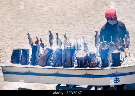 Ein Koch, der Fisch, Sardinen und Wolfsbarsch auf Spießen über einem Holzgrill im Freien kocht. Das BBQ in einem Boot befindet sich am Strand an der Costa del Sol Stockfoto