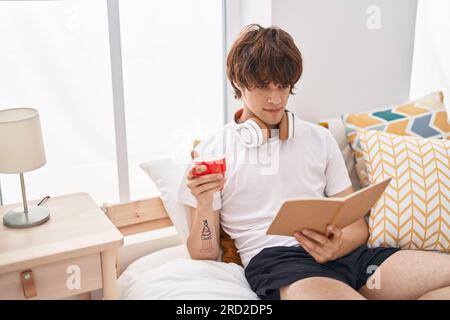 Junger blonder Mann liest Buch und trinkt Kaffee im Schlafzimmer Stockfoto