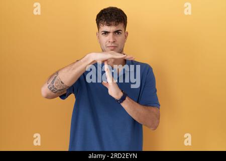 Junger hispanischer Mann, der auf gelbem Hintergrund steht und mit Händen, frustrierten und ernsten Gesichten eine Auszeit-Geste tut Stockfoto