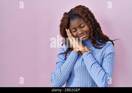 Afrikanische Frau, die über rosa Hintergrund steht, schläft müde, träumt und posiert mit den Händen zusammen, während sie mit geschlossenen Augen lächelt. Stockfoto