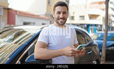 Junger hispanischer Mann mit Smartphone, der auf der Straße auf das Auto gelehnt ist Stockfoto