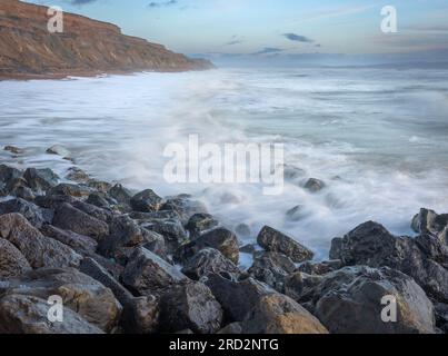 Eine frühe Meereslandschaft mit glänzenden Felsen im Vordergrund, verschwommenen Wellen über Felsen und hohen Sandsteinklippen, die in die Ferne führen Stockfoto