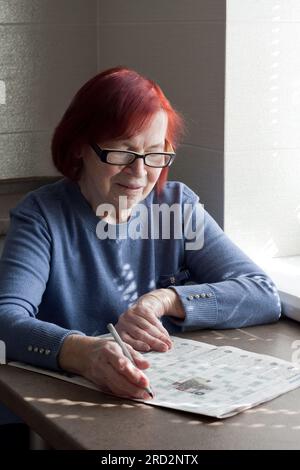 Porträt einer rothaarigen älteren Frau mit Brille, die in ihrem Haus am Tisch sitzt und Kreuzworträtsel löst. Kognitive Rehabilitationstherapie im Alter Stockfoto