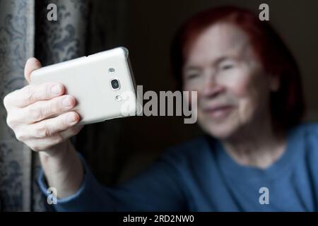Ältere, fröhliche Frau, die ihr Smartphone in der Hand hält, auf die Smartphone-Webkamera schaut, bei einem Videoanruf telefoniert, mit einem Freund aus der Ferne kommuniziert, die Anrufe entgegennimmt Stockfoto