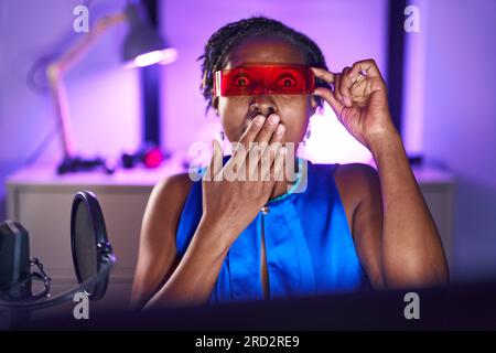 Afrikanische Frau mit Dreadlocks, die Videospiele spielt, die eine Virtual-Reality-Brille trägt, die den Mund mit der Hand verdeckt, schockiert und vor Fehlern gefürchtet ist. Überrascht Stockfoto