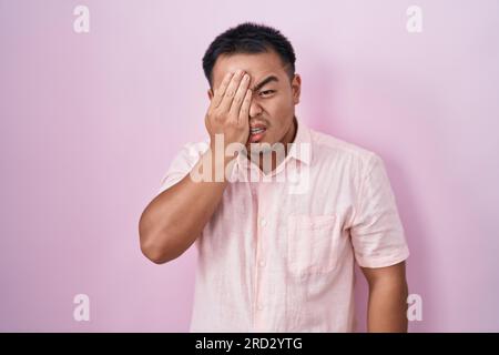 Chinesischer junger Mann, der über rosa Hintergrund steht und müde gähnt, das halbe Gesicht, Auge und Mund mit der Hand bedeckt. Das Gesicht tut weh. Stockfoto