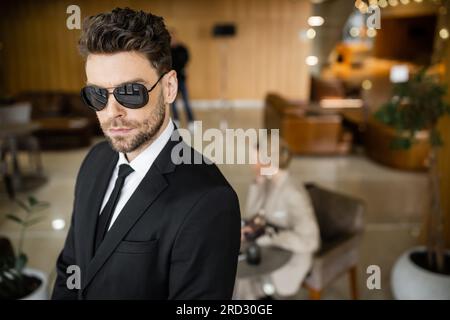 Privater Sicherheitsdienst, persönliches Sicherheitskonzept, attraktiver Leibwächter in formeller Kleidung und Sonnenbrille zum Schutz der weiblichen Klientin auf verschwommenem Rücken Stockfoto