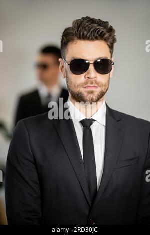 Leibwächter-Service, attraktiver Mann mit Sonnenbrille und schwarzem Anzug mit Krawatte, Hotelsicherheit, Sicherheitsmanagement, Überwachung und Wachsamkeit, uniformierter Wachmann Stockfoto
