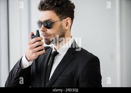Überwachung, gut aussehender Bodyguard kommuniziert über Walkie-Talkie, Mann mit Sonnenbrille und schwarzem Anzug mit Krawatte, Hotelsicherheit, Sicherheitsmanagement, un Stockfoto