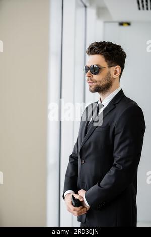 Überwachung, Bodyguard steht mit Walkie-Talkie, attraktiver Sicherheitsmann in Sonnenbrille und schwarzem Anzug mit Krawatte, Hotelsicherheitsmanagement, uniformiertes G. Stockfoto