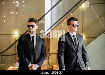 Sicherheitsmaßnahmen in Luxushotels, zwei gutaussehende Männer in formeller Kleidung und Sonnenbrille, Leibwächter im Dienst, Sicherheitsmanagement, Wachsamkeit, Anzüge und Krawatten, P Stockfoto