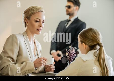 Blonde Frau hält eine Tasse Kaffee in der Hand und spricht mit einer jungen Tochter in der Nähe eines Leibwächters auf verschwommenem Hintergrund, Sicherheitsdienst, privates Sicherheitskonzept, qua Stockfoto