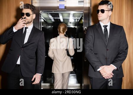Persönliches Sicherheits- und Schutzkonzept, blonde und erfolgreiche Frau mit Handtasche im Aufzug neben Leibwächtern in Anzügen und Sonnenbrillen, Luxus Stockfoto