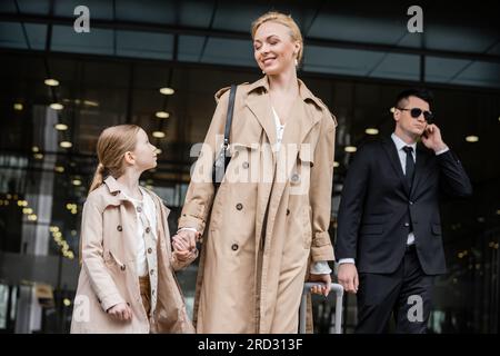 Persönliche Sicherheit, Lebensstil, blonde Mutter mit einem jungen Mädchen, das Händchen in der Nähe des Hotels hält, erfolgreiche Frau und Kind, Bodyguard im Anzug, der auf dem Fleck steht Stockfoto