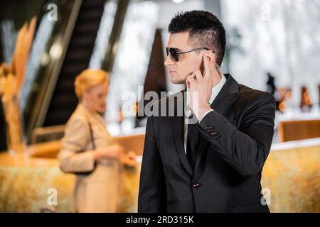 Bodyguard-Konzept, gutaussehender Mann in formeller Kleidung und Krawatte berührender Ohrhörer in der Lobby des Hotels, Sicherheit, Kommunikation, private Sicherheit, Schutz der Frau Stockfoto