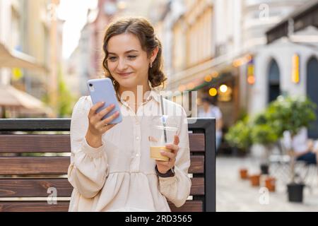 Lächelnde junge Frau, die auf dem Smartphone Text eingibt, Nachrichten beantwortet und online chattet. Sie sieht aus wie eine Social-Media-App auf dem Mobilbildschirm. Ein Touristenmädchen sitzt auf einer Bank auf der Straße der Stadt und trinkt kalten Kaffee mit Eis Stockfoto