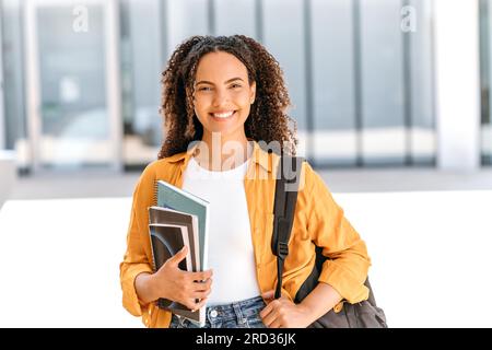Porträt einer erfolgreichen, positiven Studentin, brasilianischer oder lateinamerikanischer Nationalität, mit Rucksack, mit Büchern und Notizbüchern in der Hand, stehen in der Nähe des Universitätscampus, schauen und lächeln vor der Kamera Stockfoto