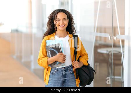 Positive, trendige Studentin. Glückliche, reizende brasilianische oder hispanische Studentin, mit einem Rucksack, mit Büchern und Notizbüchern in der Hand, steht in der Nähe des Universitätscampus, sieht und lächelt vor die Kamera Stockfoto