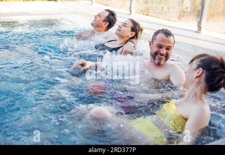 Zwei verheiratete Paare unterhalten sich und nehmen einen Außenpool. Freunde lachen und entspannen sich im wirbelnden Wasser. Junge Leute entspannen sich in einem Spa oder Hotel. Stockfoto