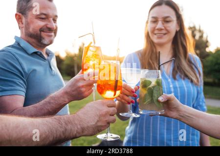 Treffen von Freunden im Hinterhof des Hauses oder in einem Café unter freiem Himmel. Junge Männer und Frauen halten Gläser mit alkoholischen Getränken im Freien. Lächeln p Stockfoto