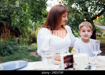 Eine Frau mittleren Alters in elegantem weißen Kleid und ein süßer Junge an einem festlichen Tisch im Freien. Großmutter und ihr Enkel feiern ihren Geburtstag im Garten oder Garten Stockfoto
