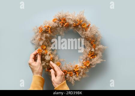Frau hält in den Händen Erntedankkranz mit orangen Blumen und trockenen natürlichen Materialien auf blauem Hintergrund. Draufsicht. Stockfoto