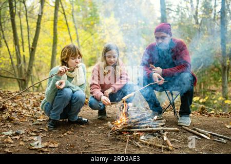 Vater und Kinder rösten Marshmallows am Lagerfeuer. Kinder haben Spaß am Lagerfeuer. Camping mit Kindern im Herbstwald. Familienfreundin Stockfoto